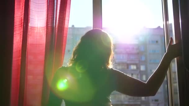幸せな若い女の子は窓に来る、それを開き、手を振り。太陽光線は、指を通過します。日の入り時刻 — ストック動画