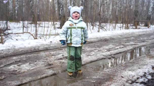 Un enfant dans le parc sautant sur des flaques d'eau. Des éclaboussures d'eau coulent sur les fêtes. Divertissement, sports de plein air. Début printemps . — Video