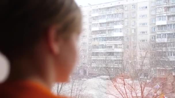 Sunny zimy den. Mladý kluk stojí u okna a dívá na padající sníh — Stock video