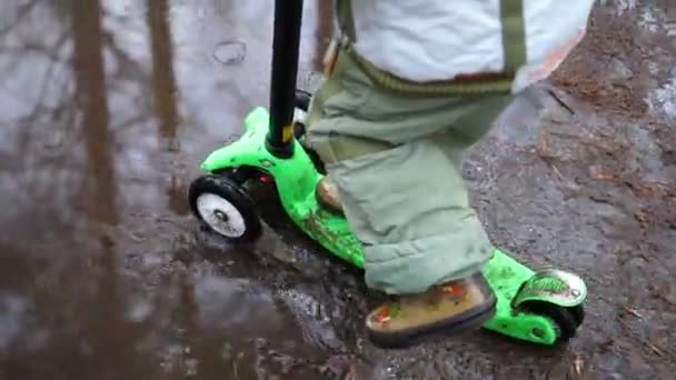 Barnet rider en skoter i parken tidigt på våren. Smältande snö, snö och pölar på asfalt. Utomhussporter — Stockvideo