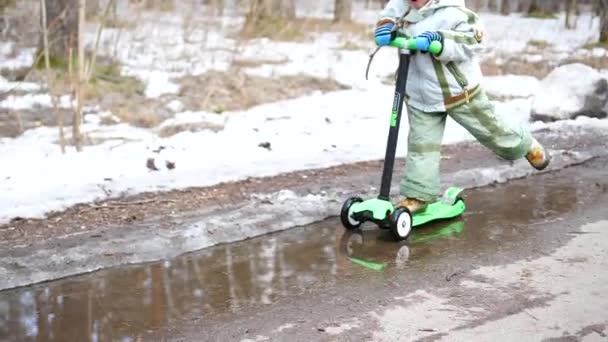 L'enfant fait du scooter dans le parc au début du printemps. Neige fondante, neige et flaques d'eau sur asphalte. Sports de plein air — Video