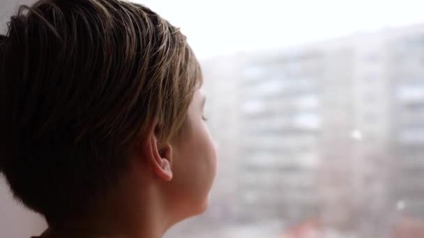 Día soleado de inviernos. Un joven se para junto a la ventana y mira la nieve que cae — Vídeos de Stock