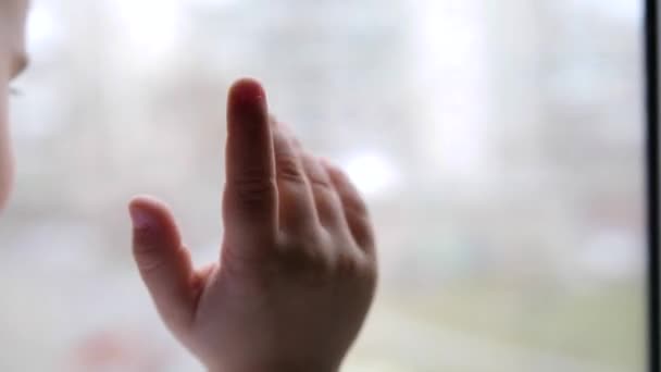 Ein Kind steht neben einem Fenster und beobachtet, wie Schnee auf die Straße fällt. die Nahaufnahme Hand am Glasfenster — Stockvideo