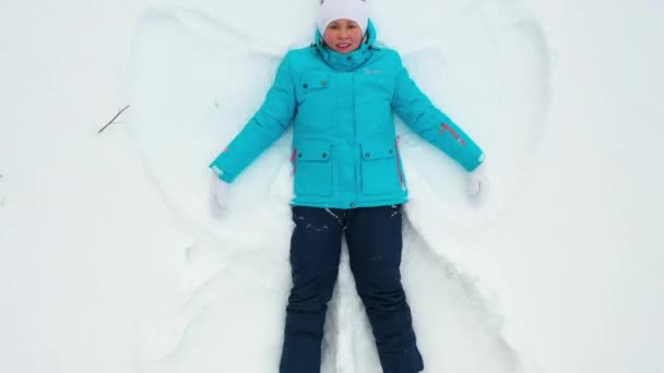 Una niña hace una figura de un ángel de la nieve en un claro en el bosque. Fotografía aérea de una chica tumbada en la nieve . — Vídeos de Stock