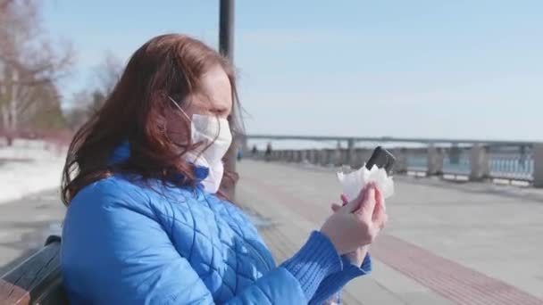 The girl wipes her mobile phone with a napkin in a protective mask on the street. Prevention and protection of health and safety of life. — Stock Video