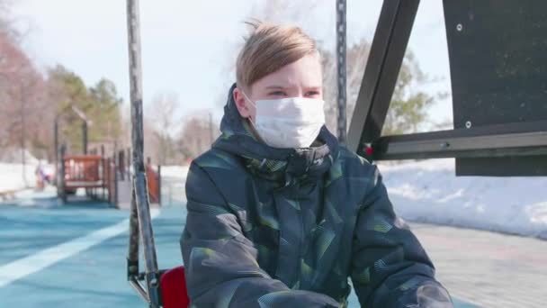 Un ragazzo è seduto su un'altalena con una maschera protettiva sul parco giochi.Prevenzione e protezione della salute e della sicurezza della vita . — Video Stock