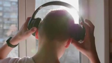Genç bir adam pencerenin yanında duruyor ve müzik dinlemek için kulaklık takıyor. Bulanık arka plan, günbatımı, gençlerin kulaklıkla müzik dinlemesi..