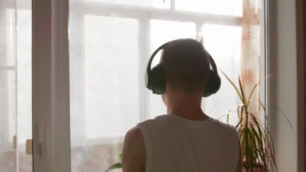 一个年轻人站在窗边，戴上耳机听音乐。背景模糊，夕阳西下，青少年在耳机里欣赏音乐. — 图库视频影像