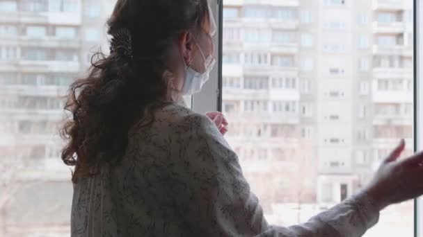 Девушка в защитной маске сидит дома в самоизоляции. Девушка смотрит в открытое окно. Профилактика и защита здоровья и безопасности жизни . — стоковое видео