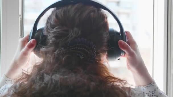 Una joven va a la ventana y se pone los auriculares para escuchar música. Una mujer disfruta de la melodía en sus auriculares . — Vídeos de Stock
