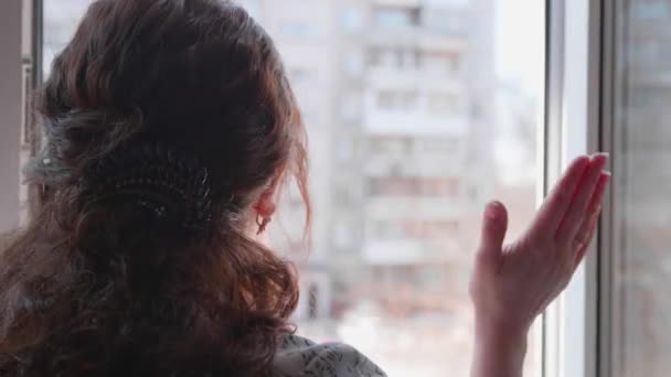 護身用のマスクの少女が自宅で自立して座っている。コロナウイルスと戦っている人々を支援するために窓の中で少女がスラム. — ストック動画