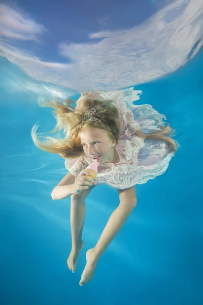 Girl in white dress eating ice cream underwater. Young beautiful girl poses underwater in the pool.