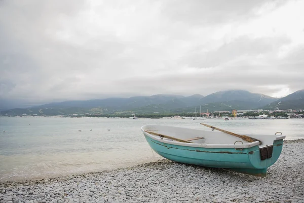 side view of small wooden fishing azure boat on pebble coast black sea beach in bad weather on dramatic sea, sky, mountain and town background, horizontal stock photo image