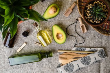 Çevre dostu mutfak eşyaları. Ahşap çatal bıçak takımı, cam süs, seramik kapta bir çiçek, doğal yağlar ve sarı bir duvara karşı keten masa örtüsü üzerinde dilimlenmiş bir avokado. Sıfır atık ve plastikten