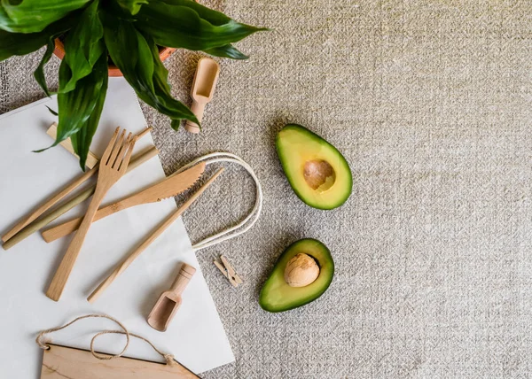 Set of eco friendly kitchen items. wooden cutlery, glassware, a flower in a ceramic pot, natural oils and a sliced avocado on a linen tablecloth against a yellow wall. Zero waste and plastic free