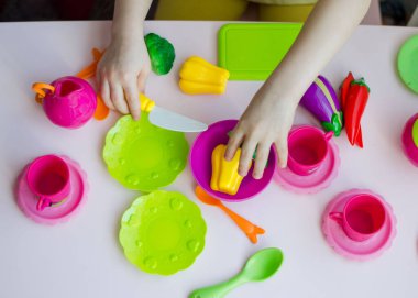 Küçük kız plastik bulaşıklar ve sebze oyuncaklarıyla aşçılık oynuyor. Brokoli, paprica gibi renkli plastik sebzeli çocukların elleri. Yaklaş, seçici odaklan.