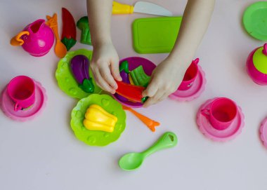 Küçük kız plastik bulaşıklar ve sebze oyuncaklarıyla aşçılık oynuyor. Brokoli, paprica gibi renkli plastik sebzeli çocukların elleri. Yaklaş, seçici odaklan.