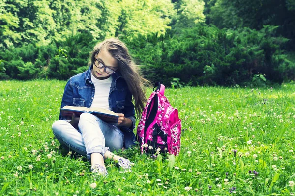 公園でバックパックを持つ草の本を読んでジーンズ シャツの美しいブロンド女子高生女の子 — ストック写真