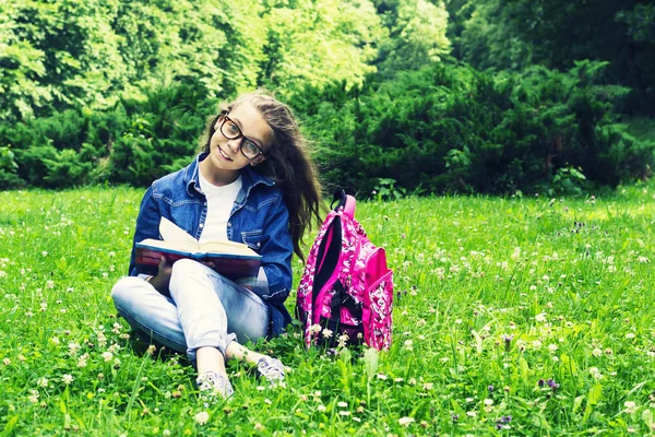 Vacker blond Skolflicka tjej i jeans skjorta en bok på gräset med en ryggsäck i parken — Stockfoto