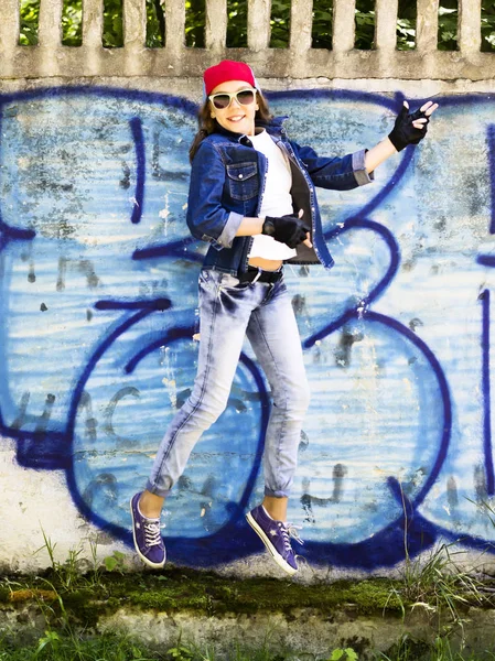 Menina adolescente loira bonito em um boné de beisebol e camisa jeans saltando contra um fundo de parede de pedra. Salto do quadril , — Fotografia de Stock