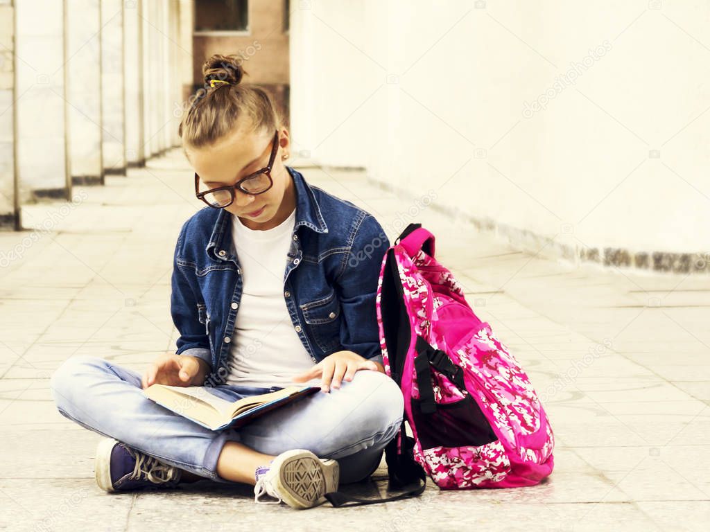 Cute fair-haired girl schoolgirl reading a book while sitting near school. Education.