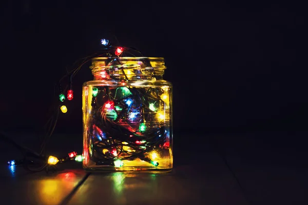 A glowing garland in a glass jar and Christmas decorations on a dark wooden background. New Year, Christmas postcard. Copy space. — Stock Photo, Image