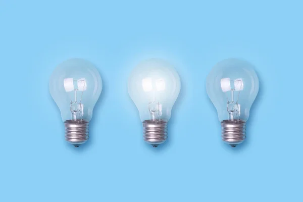 Una bombilla luminosa y dos no luminosas sobre un fondo azul. El concepto de nuevas ideas. Negocios. Creación . — Foto de Stock