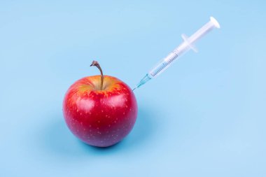 Mavi arka planda kırmızı bir elmaya şırınga enjekte edilmiş. Genetik modifiye edilmiş gıdalar. Ben...