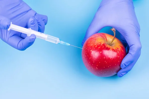 Wstrzyknięcie do jabłka. Ręka w rękawiczce medycznej ze strzykawką na niebieskim tle. Genetycznie zmodyfikowana żywność. — Zdjęcie stockowe