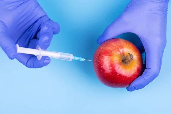 Wstrzyknięcie do jabłka. Ręka w rękawiczce medycznej ze strzykawką na niebieskim tle. Genetycznie zmodyfikowana żywność. — Zdjęcie stockowe