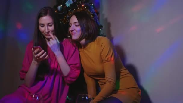 Weibliches Paar sitzt mit Weingläsern am Weihnachtsbaum und schaut Videos — Stockvideo