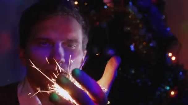 Jonge man bedrijf sparkler zitten in de buurt van een kerstboom — Stockvideo