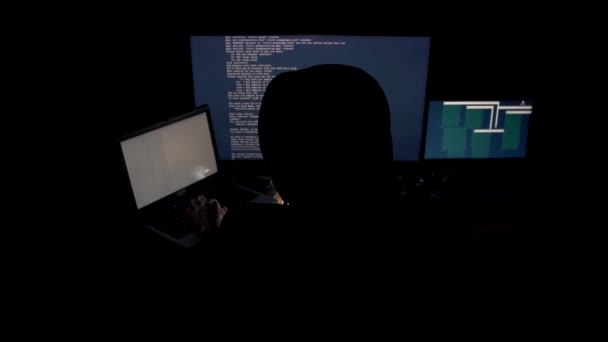 Хакер в капюшоне взламывает код с помощью компьютеров в темной комнате — стоковое видео