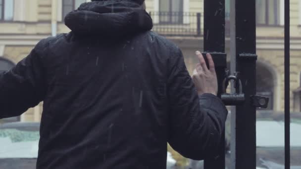 Mladý muž stál a čekal poblíž železné brány pod sněhem — Stock video