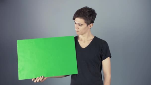 Hombre joven feliz en camisa negra sosteniendo el cartel verde de la hoja de la llave fondo gris — Vídeo de stock