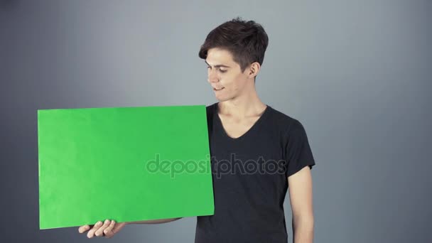 Leende ung man i svart tröja holding grön nyckel ark affisch grå bakgrund — Stockvideo