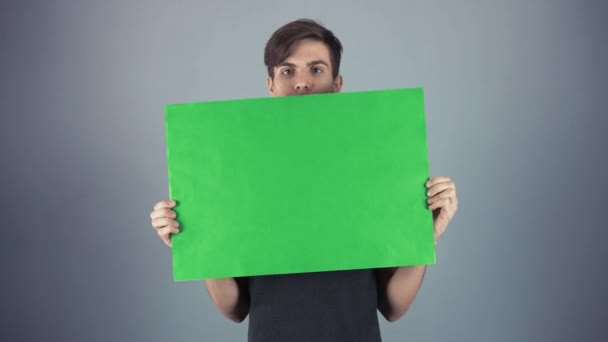Szczęśliwy młody mężczyzna w czarnej koszuli przytrzymując zielony klucz arkusz plakat szary tło — Wideo stockowe
