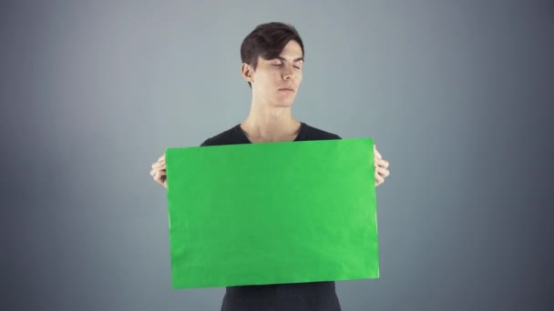 Descontente Jovem homem em camisa preta segurando verde folha-chave cartaz cinza fundo — Vídeo de Stock