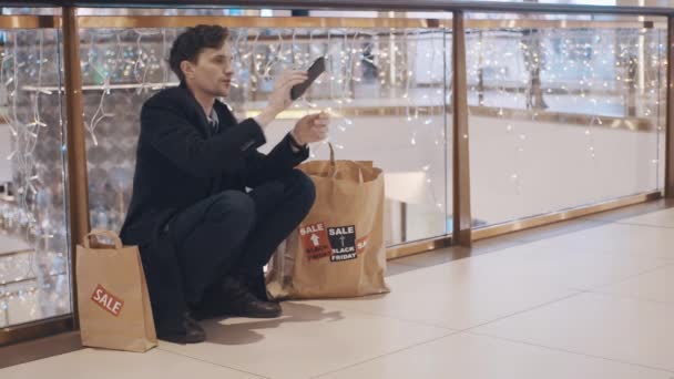Decepcionado joven sentado en el suelo en el centro comercial sin dinero después de las ventas del Viernes Negro — Vídeo de stock