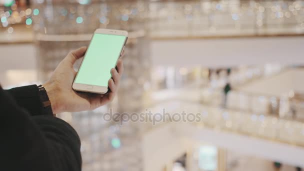 Manos masculinas usando smartphone con pantalla verde en el centro comercial. Decoraciones navideñas en segundo plano — Vídeo de stock