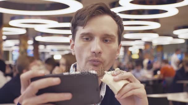 Los hombres jóvenes que usan teléfonos inteligentes comen Shawarma roll en el patio de comidas en el centro comercial. Retrato — Vídeos de Stock