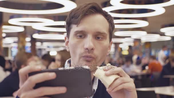 Los hombres jóvenes que usan teléfonos inteligentes comen Shawarma roll en el patio de comidas en el centro comercial. Retrato — Vídeo de stock