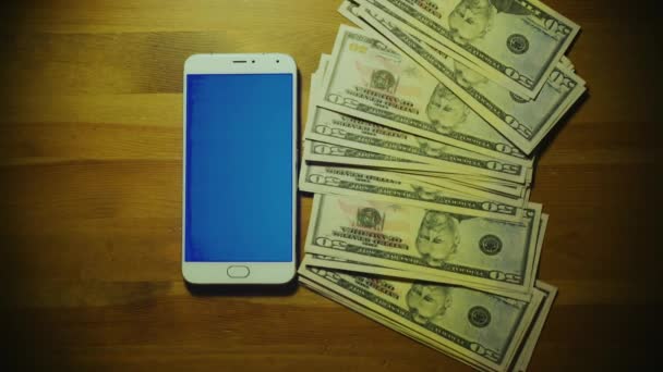 Male hand throws pile of money on srceen of white smartphone with blue key on wooden surface — Stock Video