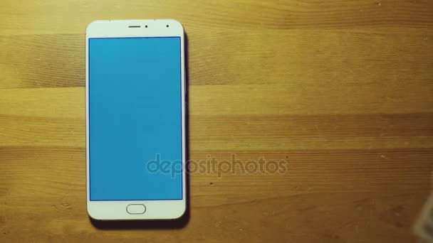 Male hand throws pile of dollars on srceen of white smartphone with blue key on wooden surface — Stock Video