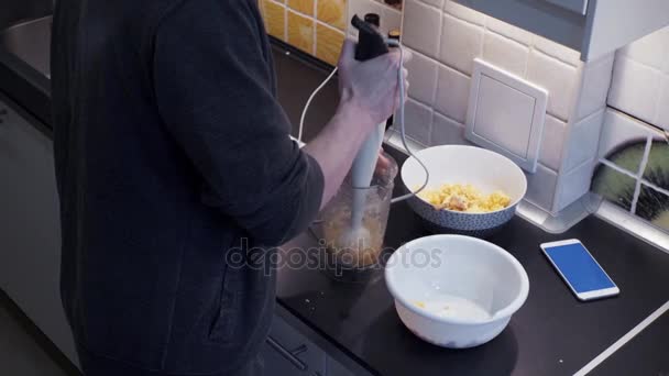 Молодой человек с блендером готовит овощную еду на домашней кухне и смартфоне — стоковое видео