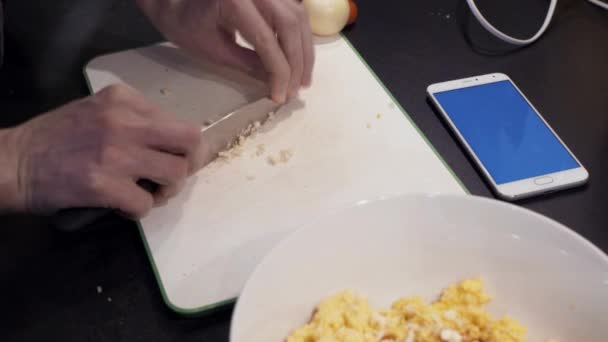 Manos de joven cortando ajo en la cocina casera. smartphone — Vídeos de Stock