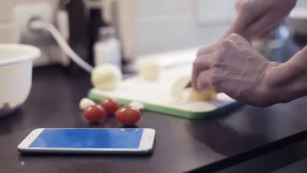 Männliche Hände schneiden in der Küche Zwiebeln und bereiten mit dem Smartphone Gemüsemahlzeit zu — Stockvideo