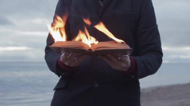 Jovem com livro em chamas na mão em pé na costa nuvens tempestade no fundo — Vídeo de Stock