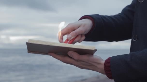 Jovem Igniting livro na mão de pé na costa nuvens tempestade no fundo — Vídeo de Stock