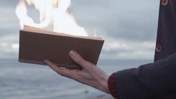 Jovem com livro em chamas na mão em pé na costa nuvens tempestade no fundo — Vídeo de Stock